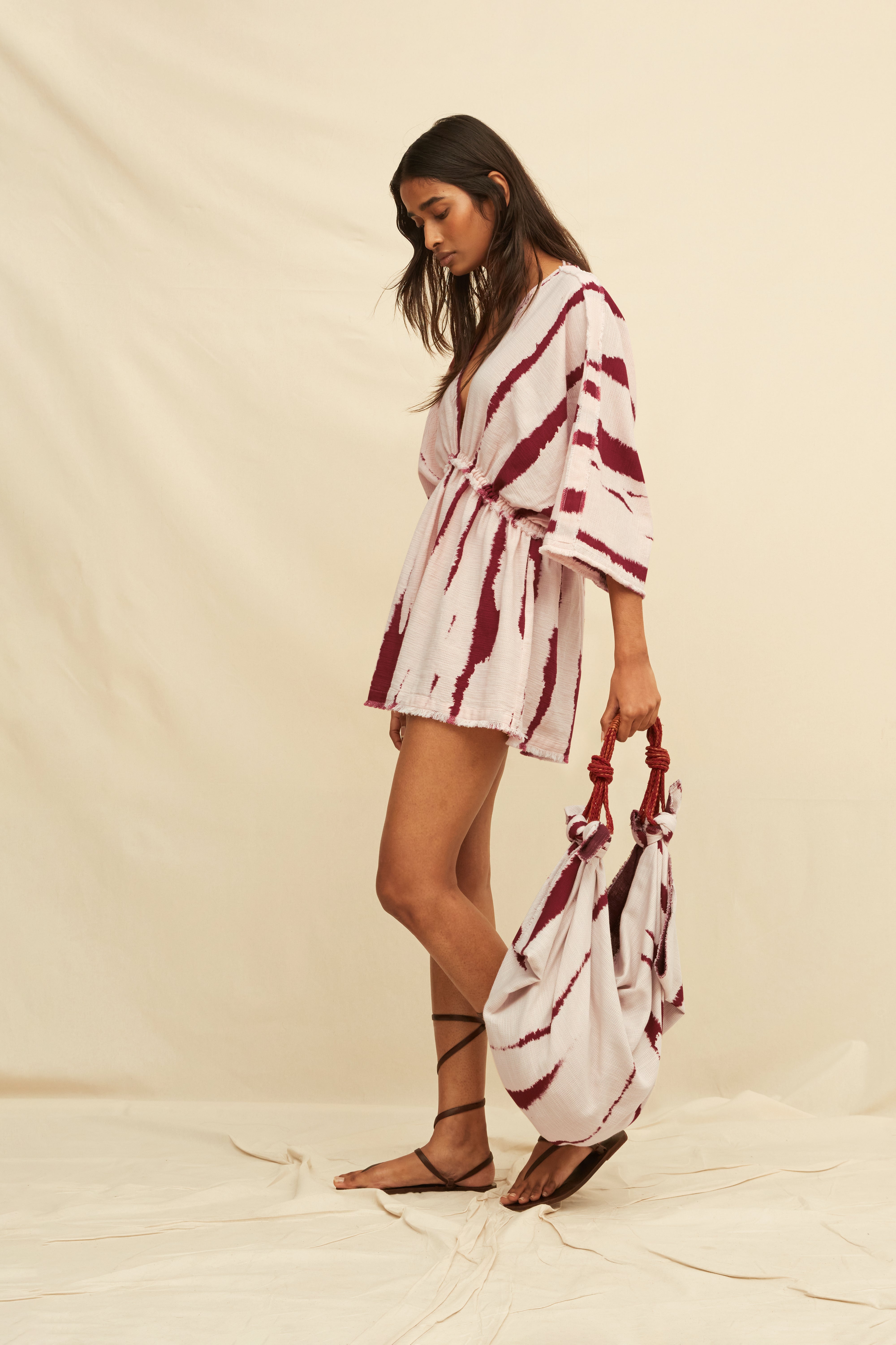 Woman wearing a tie-dye kaftan dress holding a matching bag and gladiator sandals, on a beige background