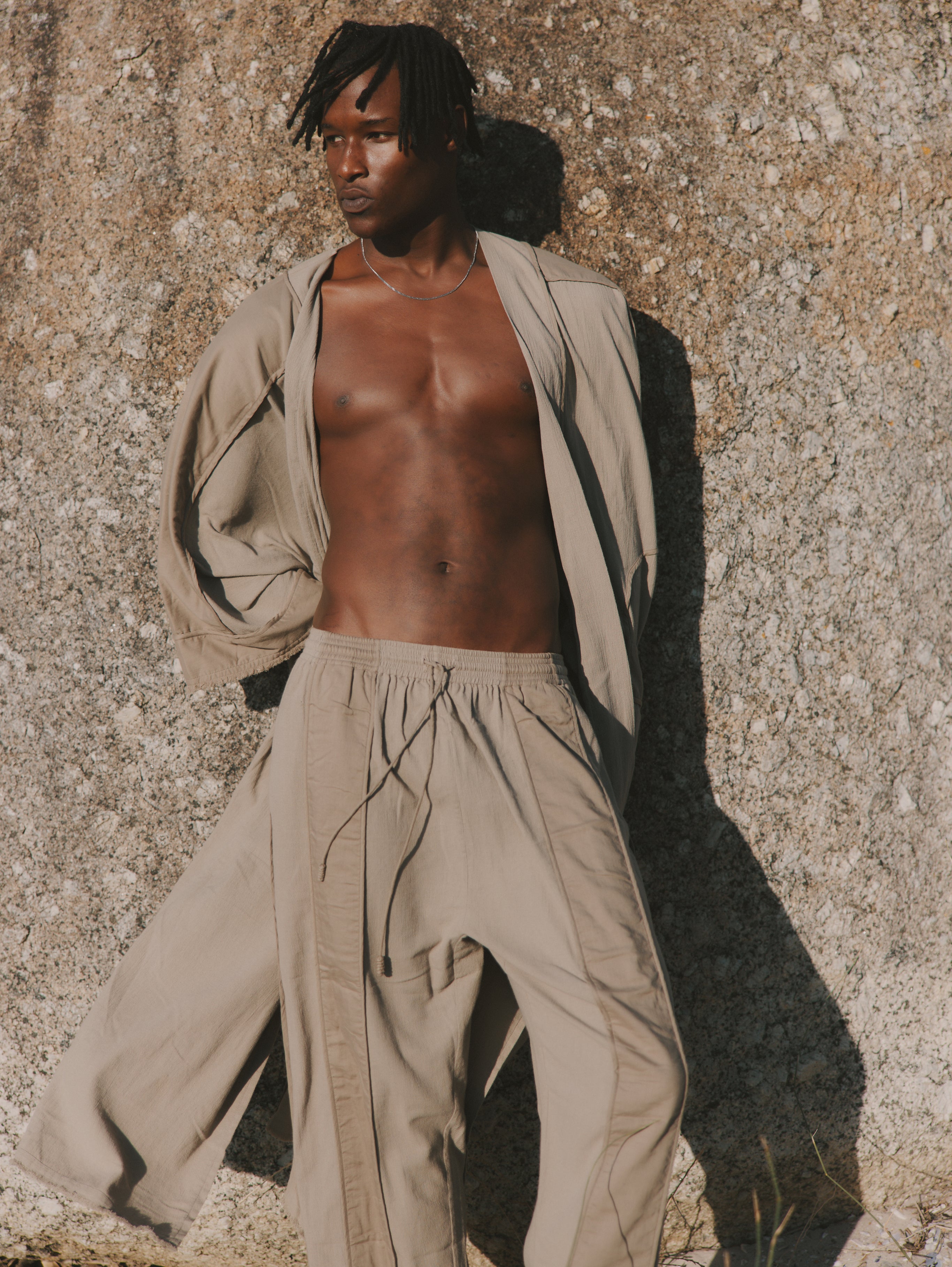 Man wearing trousers and long cover-up standing against a wall