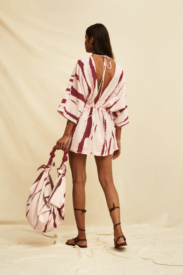 Woman wearing a tie-dye kaftan dress holding a matching bag and gladiator sandals, on a beige background