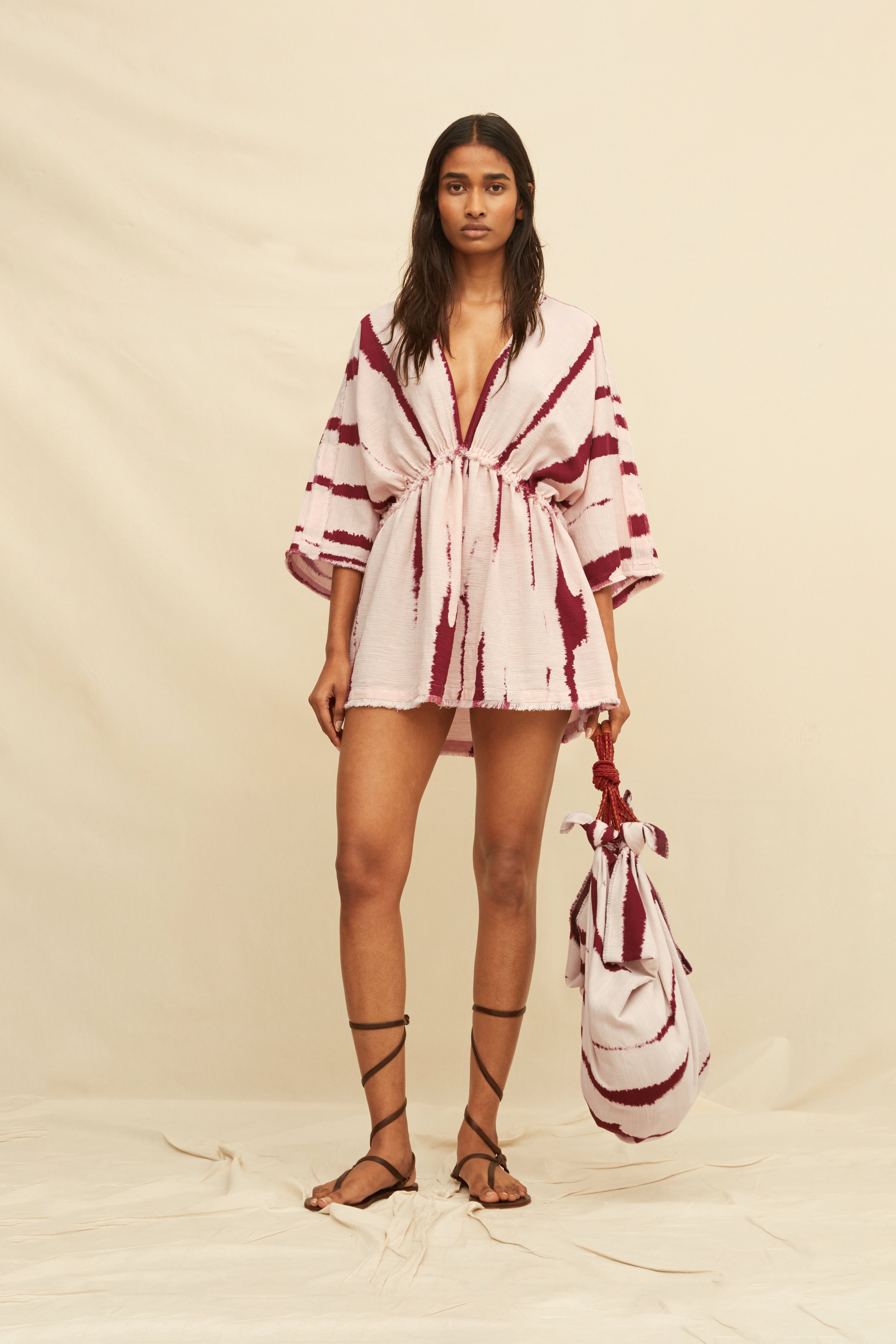 Woman wearing a tie-dye kaftan dress holding a matching bag and gladiator sandals, on a beige background