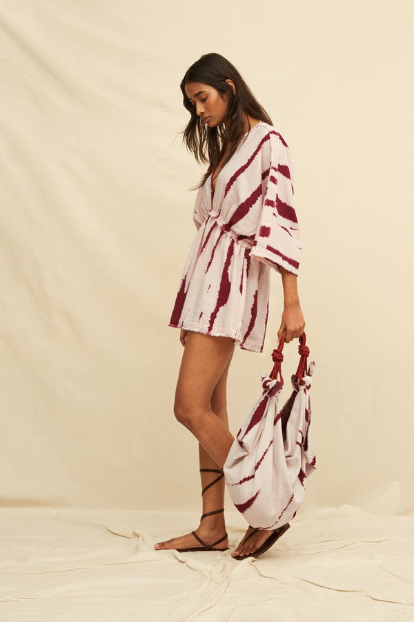 Woman wearing a tie-dye kaftan dress holding a matching bag and gladiator sandals, on a beige background