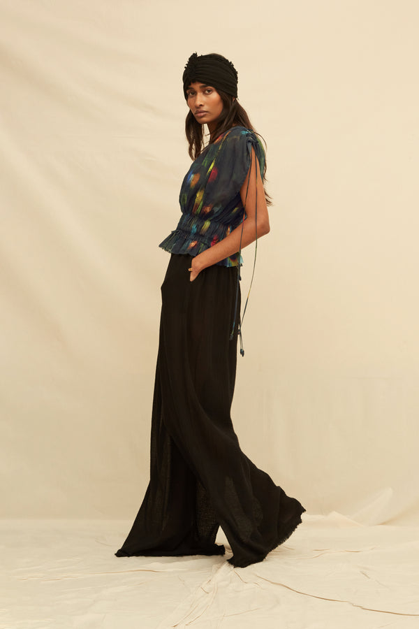 Woman wearing a black blouse with abstract painterly pattern, paired with black trousers and a black turban, on a beige background