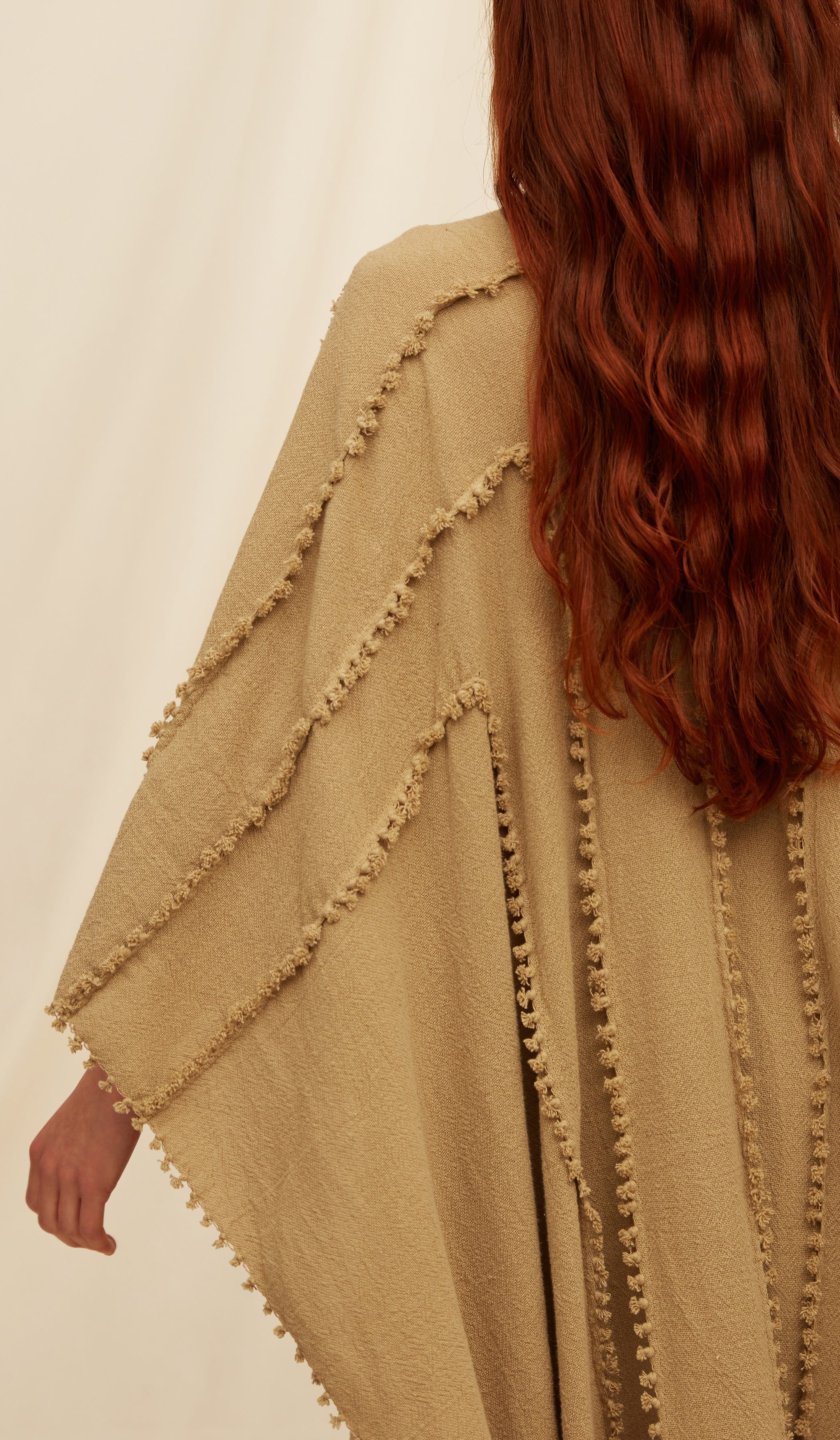 A close-up of a person wearing Yalchen Knots Sepia cape with decorative knots trim, shown from the back against a plain light background.